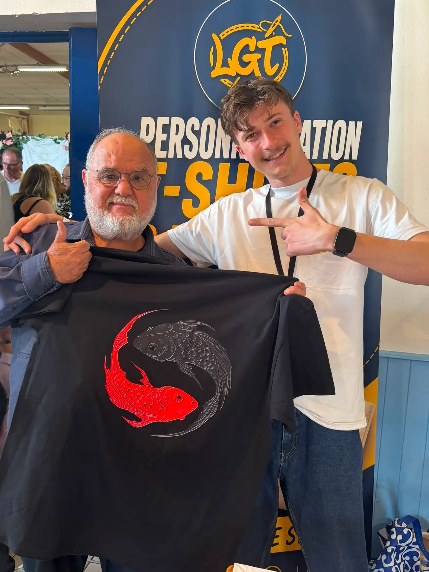 Sur le stand de l'association, un visiteur et un bénévole tiennent ensemble le t-shirt noir qui vient d'être imprimé, orné de deux carpes koï rouge et grise.